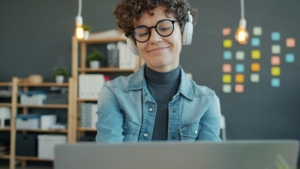 A person with short curly hair and thick rimmed black glasses sits smiling at their laptop with a grid of post it notes behind them.