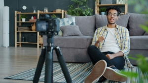 A man with short afro texture hair is sitting on his floor leaning against a sofa. He is looking at a camera set up on a tripod.