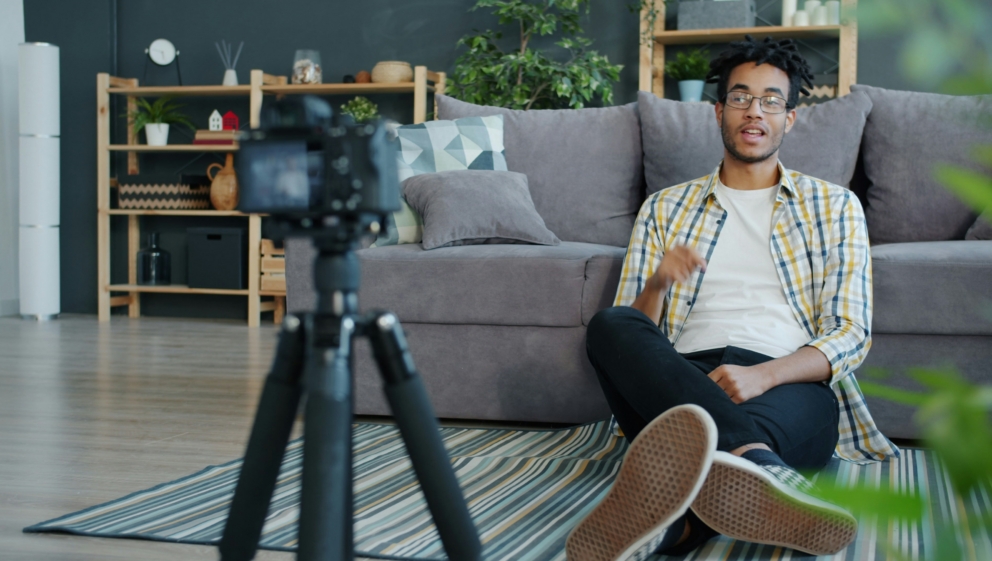 A man with short afro texture hair is sitting on his floor leaning against a sofa. He is looking at a camera set up on a tripod.