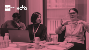 Pink-toned image showing people sat around a desk, using laptops and talking.