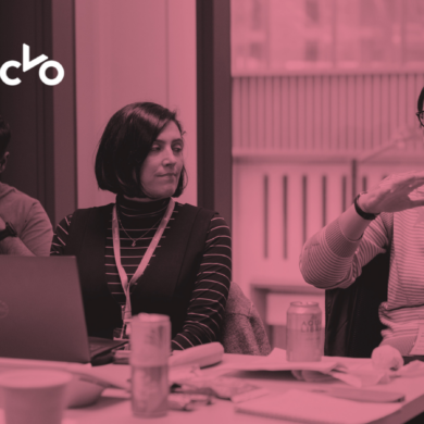 Pink-toned image showing people sat around a desk, using laptops and talking.
