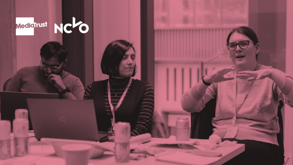 Pink-toned image showing people sat around a desk, using laptops and talking.