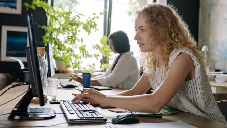A person, sat a desk, looking at a computer screen as they type on a keyboard. Behind them, another person is doing the same.