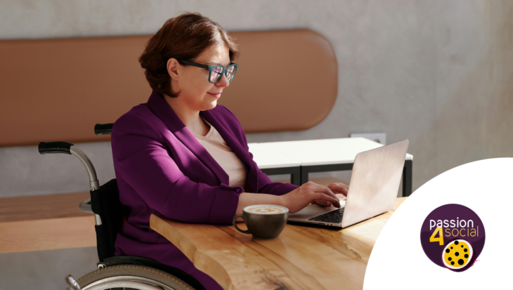 A woman with short brown hair and black glasses sits in a wheelchair at a desk with a laptop. She is wearing a dark purple blazer, and is typing on her laptop