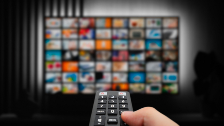 A close-up of a handing holding a remote control, pointing at a TV screen showing multiple channels.