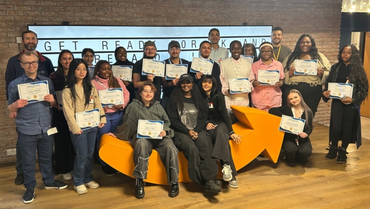 A large group of people, at the Amazon offices, posing as they hold up certificates. Some are sat on a large orange arrow.