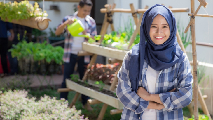 A person smiling and posing in front of an urban garden. Behind them, another person is watering the plants.