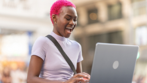 A person with bright pink hair, smiling widely as they use a laptop