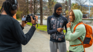 Three young people outside. One is holding a mic, interviewing another, whilst a third films.