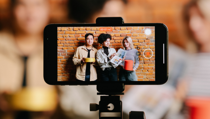 A mobile phone on a tripod, filming 3 people who are each holding an object.