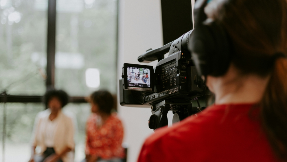 Man in red shirt facing away from the camera holding his own large filming camera pointed towards two women in the distance